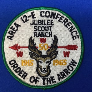 Puede incluir: Un parche bordado blanco y verde con el texto "Area 12-E Conference Jubilee Scout Ranch Order of the Arrow 1915-1965" y un ciervo con cuernos y una flecha.