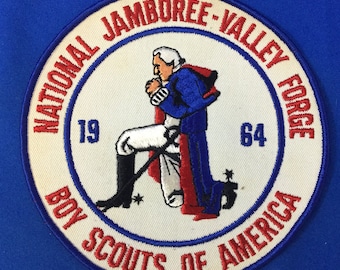 Pode incluir: Um remendo branco redondo bordado com detalhes vermelhos e azuis. Apresenta uma figura ajoelhada com um casaco azul e calças brancas, com o texto "National Jamboree - Valley Forge 1964 Boy Scouts of America".