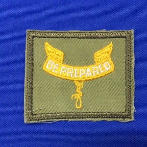 Könnte beinhalten: Ein grüner Stoff-Patch mit einem gelben gestickten Rand und einem gelben gestickten Banner mit den Worten "BE PREPARED" in Weiß.