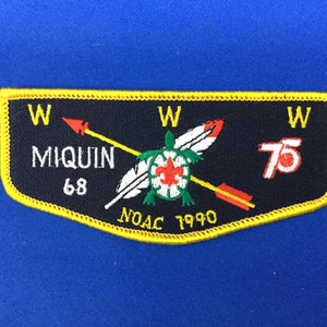 Puede incluir: Un parche rectangular con fondo negro y borde amarillo. El parche presenta el texto "MIQUIN 68" y "NOAC 1990". Una tortuga con una flor de lis está en el centro, con flechas y plumas. El número "75" está a la derecha.