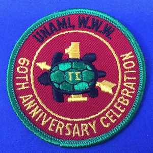 Puede incluir: Un parche bordado en rojo, verde y dorado con una tortuga y el texto "60th Anniversary Celebration W.H.A.M.L. W.J.W.W."