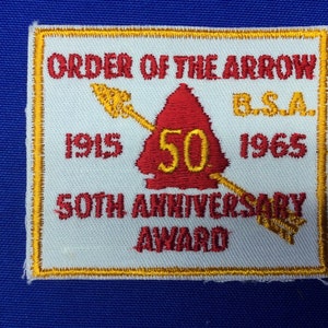 Puede incluir: Un parche blanco con borde dorado, con el texto "ORDER OF THE ARROW BSA 1915 1965 50TH ANNIVERSARY AWARD". En el centro, una punta de flecha roja con el número 50 en amarillo.
