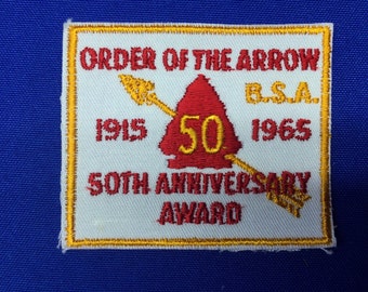 May include: A white patch with a gold border, featuring the text "ORDER OF THE ARROW BSA 1915 1965 50TH ANNIVERSARY AWARD". A red arrowhead with the number 50 in yellow is in the center.