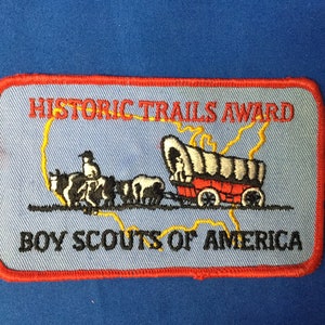 May include: A blue and red embroidered patch with the text "Historic Trails Award" and "Boy Scouts of America". The patch features a wagon train with a covered wagon, oxen, and a person riding a horse.