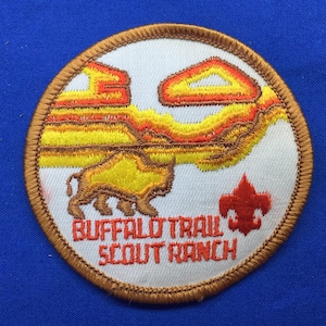 Pode incluir: Um remendo redondo bordado de cor marrom com fundo branco. O remendo apresenta uma silhueta de búfalo amarela e laranja com uma cordilheira de montanhas ao fundo. O texto "Buffalo Trail Scout Ranch" está escrito em vermelho abaixo do búfalo.