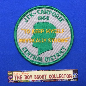 Może przedstawiać: Naszywka haftowana w kolorze zielonym i szarym z tekstem "JFK-Campore 1964" i "To Keep Myself Physically Strong" w kolorze żółtym. Naszywka zawiera również tekst "Central District" w kolorze zielonym.