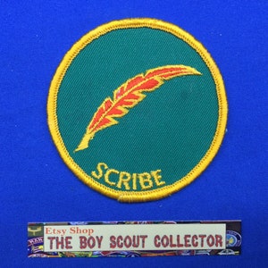 Op de afbeelding: Een groene en gele geborduurde scoutingbadge met een rode veerpen en het woord "SCRIBE" in gele letters.