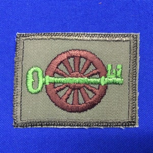 Op de afbeelding: Geborduurde patch met een bruin wagenwiel en een groen sleutelontwerp. De sleutel loopt horizontaal door het midden van het wiel. De patch heeft een grijze achtergrond en een donkergrijze rand. De patch staat op een blauwe achtergrond.