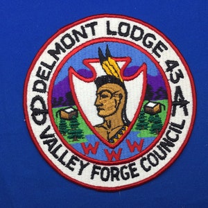 Könnte beinhalten: Ein gestickter Aufnäher in Rot, Weiß und Blau mit dem Text "Delmont Lodge 43 Valley Forge Council" am Rand. Die Mitte des Aufnähers zeigt ein Profil eines amerikanischen Ureinwohners mit einem Federkopfschmuck und einer weißen Pfeilspitze.