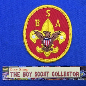 May include: A red and yellow Boy Scout of America patch with a gold eagle and a fleur-de-lis design. The patch has the letters "BSA" and "S" in yellow.