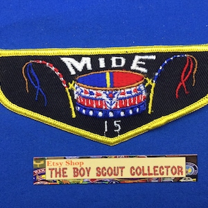 Puede incluir: Un parche de Boy Scout bordado en negro y amarillo con un tambor rojo, blanco y azul y la palabra "MIDE" encima. El número "15" está debajo del tambor.
