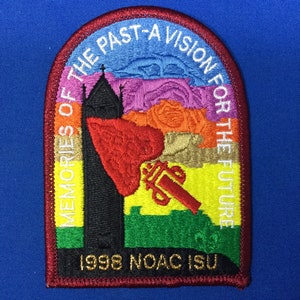 Puede incluir: Un parche bordado en rojo, blanco y dorado con el texto "Memories of the Past - A Vision for the Future" y "1998 NOAC ISU". El parche presenta una imagen estilizada de una iglesia y una antorcha.