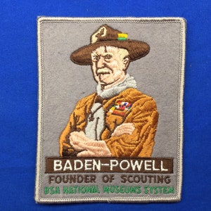 Peut inclure: Écusson brodé représentant Baden-Powell, fondateur du scoutisme. Il porte un chapeau et une veste marron. Le texte indique "Baden-Powell, Founder of Scouting, BSA National Museums System."