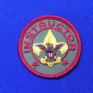 May include: A red and green embroidered patch with the word "INSTRUCTOR" around the edge. In the center is a yellow and brown embroidered scout emblem.