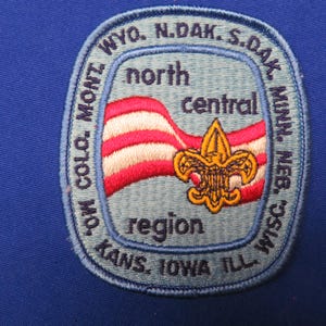 Puede incluir: Parche bordado con fondo azul claro y borde azul oscuro. El parche presenta un diseño de bandera a rayas rojas y blancas, un lirio dorado y el texto "north central region" y los nombres de varios estados.