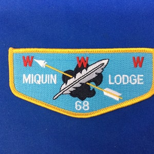 Puede incluir: Un parche bordado azul y amarillo con el texto "W Miquin Lodge W" y el número "68".