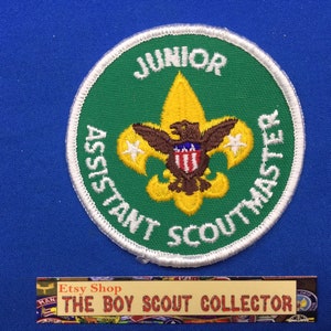 May include: A green and white embroidered patch with the text "Junior Assistant Scoutmaster" and a yellow scout emblem with a brown eagle and a shield with red and white stripes.