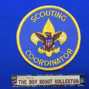 Op de afbeelding: Een geborduurde patch in blauw en goud met de tekst "SCOUTING COORDINATOR" en een gouden lelie met een adelaar en een schild.