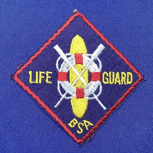 May include: A blue diamond-shaped patch with red stitching. The patch features a white life preserver with a red stripe, two crossed oars, and a yellow canoe. The text "LIFE GUARD BSA" is embroidered on the patch.