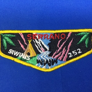 Puede incluir: Parche bordado con borde amarillo y fondo azul. El parche presenta la palabra "SERRANO" en rojo, "SIWINIS" en blanco y el número "252". El diseño incluye un tipi, una cascada y detalles en rosa y verde.