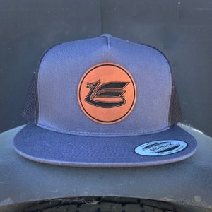 May include: A gray and black trucker hat with a brown leather patch featuring a stylized black dragon logo. The patch is circular and has a white outline.