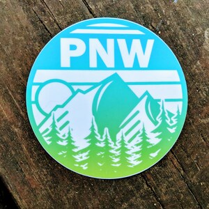 May include: A round sticker with a blue and green gradient background. The sticker features a white mountain range with a sun and the text "PNW" in white.