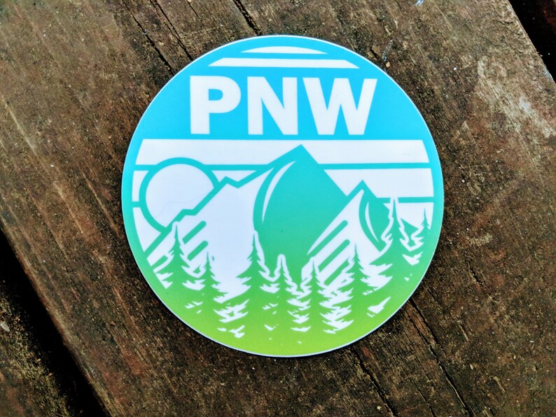 May include: A round sticker with a blue and green gradient background. The sticker features a white mountain range with a sun and the text "PNW" in white.