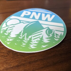 May include: A round sticker with a blue and green gradient background. The sticker features a white silhouette of a mountain range with trees in the foreground. The text "PNW" is printed in white at the top of the sticker.
