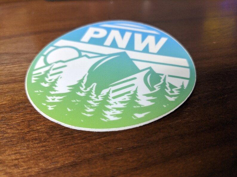 May include: A round sticker with a blue and green gradient background. The sticker features a white silhouette of a mountain range with trees in the foreground. The text "PNW" is printed in white at the top of the sticker.