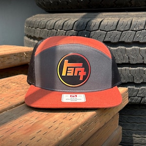 May include: A five-panel trucker hat featuring a rust-orange brim and back panels, a dark gray front panel, and black mesh side panels. A circular patch on the front displays a stylized orange and red logo. A white Richardson brand label is on the brim.