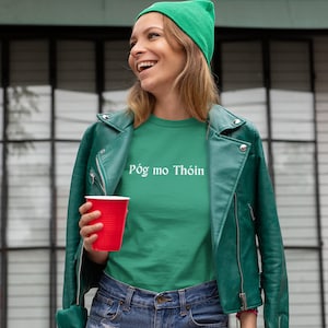 May include: A woman wearing a green beanie, a green t-shirt with the text "Póg mo Thóin" in white, a green leather jacket, and denim shorts. She is holding a red plastic cup.