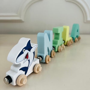 May include: A wooden toy train with a shark design on the front engine car. The train is made up of five cars, each with a different letter spelling out the name "NILA". The cars are painted in shades of blue, green, and yellow.