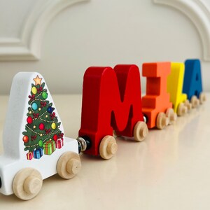 May include: A wooden toy train with colorful letters spelling out the name "MIA". The first car is white with a Christmas tree design and the remaining cars are red, orange, yellow, and blue.