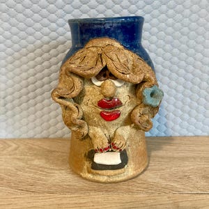 May include: A whimsical ceramic vase shaped like a woman's face. The vase features a blue top, tan face with red lips, and a blue flower. The woman's face has tan hair and is holding a black and white object.