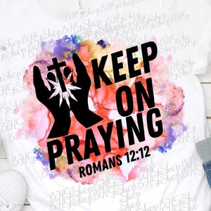 May include: White t-shirt with a watercolor background and black text that reads "KEEP ON PRAYING" with the scripture reference "ROMANS 12:12". The design also includes a pair of hands with a cross in the center.