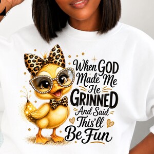 May include: A white long-sleeve sweatshirt featuring a graphic of a yellow duckling wearing leopard print bows and jewelled glasses. The text reads, When GOD Made Me He GRINNED And Said This'll Be Fun, accompanied by gold glitter accents and sparkles.