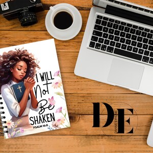 May include: A white spiral notebook with a colorful illustration of a woman with her hands clasped in prayer. The text "I WILL NOT BE SHAKEN" is printed in black with the scripture reference "PSALM 16:8" below. The background features pink and purple flowers.