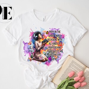 May include: White t-shirt with a watercolor design of a woman kneeling with a book and butterflies. The text "Pray in the Spirit on all occasions with all kinds of prayers and requests Ephesians 6:18" is printed on the shirt.