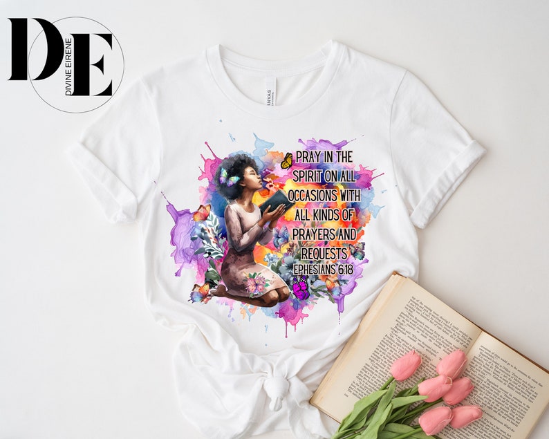 May include: White t-shirt with a watercolor design of a woman kneeling with a book and butterflies. The text "Pray in the Spirit on all occasions with all kinds of prayers and requests Ephesians 6:18" is printed on the shirt.
