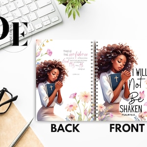May include: A pink and white floral design with a black woman holding a bible. The text on the front of the journal reads "I will not be shaken Psalm 16:8". The text on the back of the journal reads "This is the confidence that we have in approaching God: that if we ask anything according to His will, He hears us."