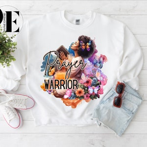 May include: White sweatshirt with a graphic print of a woman holding a book with the words "Prayer Warrior" in black text. The graphic is surrounded by colorful flowers and butterflies.