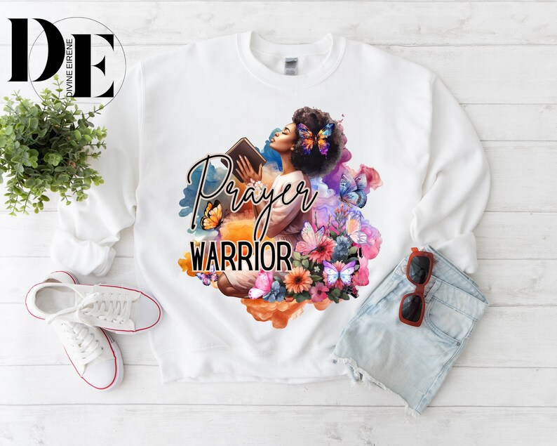 May include: White sweatshirt with a graphic print of a woman holding a book with the words "Prayer Warrior" in black text. The graphic is surrounded by colorful flowers and butterflies.