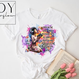 May include: A white t-shirt with a watercolor design of a woman kneeling with a book and butterflies. The text "Pray in the spirit on all occasions with all kinds of prayers and requests Ephesians 6:18" is printed on the shirt.
