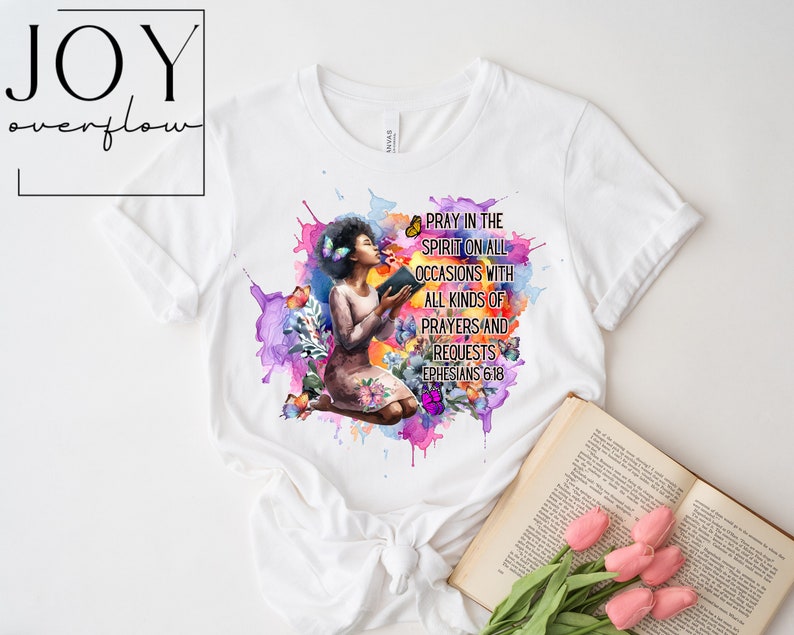 May include: A white t-shirt with a watercolor design of a woman kneeling with a book and butterflies. The text "Pray in the spirit on all occasions with all kinds of prayers and requests Ephesians 6:18" is printed on the shirt.