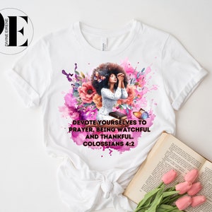 May include: White t-shirt with a watercolor design of a woman praying with flowers and butterflies. The text on the shirt reads "Devote yourselves to prayer, being watchful and thankful. Colossians 4:2"