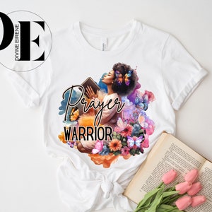 May include: White t-shirt with a colorful graphic design featuring a woman holding a book, butterflies, and flowers. The text "Prayer Warrior" is printed on the shirt.