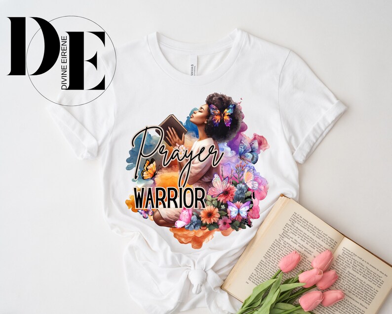 May include: White t-shirt with a colorful graphic design featuring a woman holding a book, butterflies, and flowers. The text "Prayer Warrior" is printed on the shirt.
