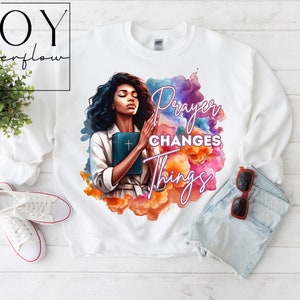 May include: White sweatshirt with a colorful watercolor design featuring a woman holding a bible and the text "Prayer Changes Things".