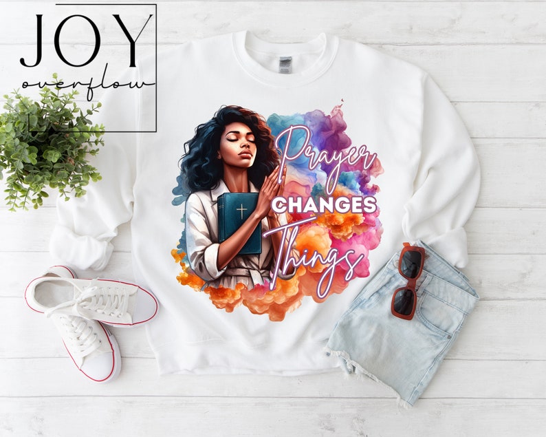 May include: White sweatshirt with a colorful watercolor design featuring a woman holding a bible and the text "Prayer Changes Things".