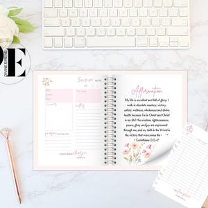 May include: A pink and white journal with a floral design on the cover. The journal has a spiral binding and includes sections for scripture, notes, prayer, and affirmations. The affirmation section includes the text "My life is excellent and full of glory: I walk, in absolute mastery, victory, safety, wellness, wholeness and divine health because I'm in Christ and Christ is my life! His wisdom, righteousness, peace, glory and joy are expressed through me, and my faith in the Word is the victory that overcomes the... 1 Corinthians 2:12-14".